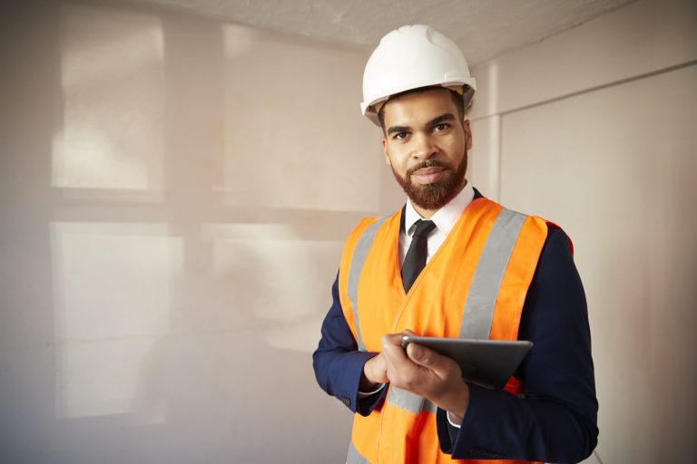 Portrait Of Surveyor In Hard Hat And High Visibility Jacket With Digital Tablet Carrying Out House Inspection